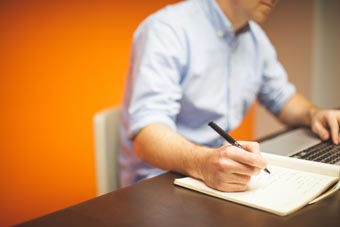 A man at a desk with one hand on his keyboard and the other holding a pen to a pad of paper
